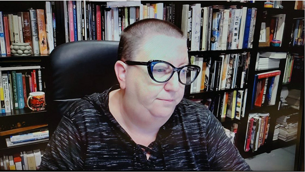 A photograph of a woman wearing glasses, looking away from the camera and books on shelves in background