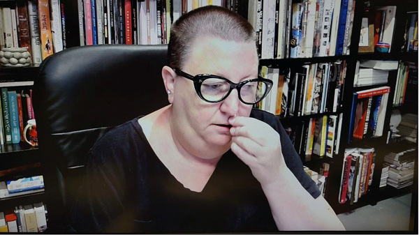 A photograph of a woman wearing glasses conducting a Covid test in her nose, looking away from the camera and books on shelves in background