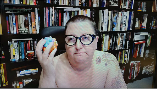 A photograph of a woman wearing glasses, holding a dog toy, looking away from the camera and books on shelves in background