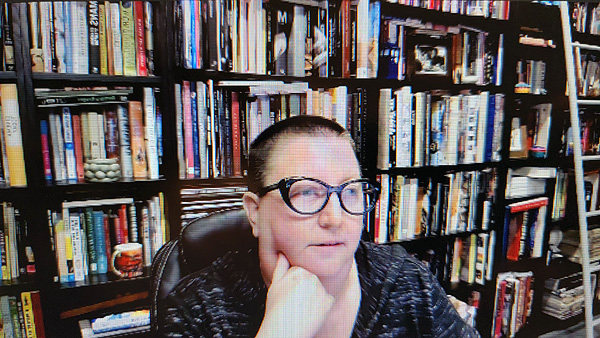 A photograph of a woman wearing glasses, her right hand resting on her chin, looking away from the camera and books on shelves in background