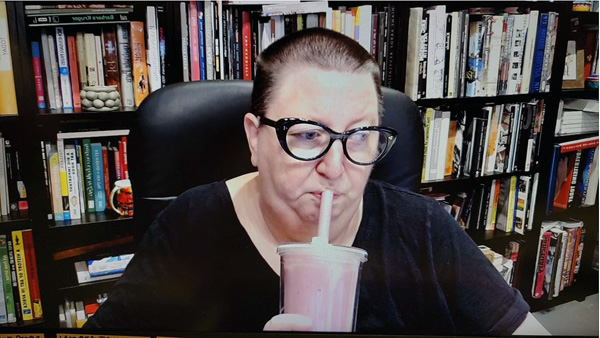 A photograph of a woman wearing glasses, sipping on a straw in a cup, looking away from the camera and books on shelves in background