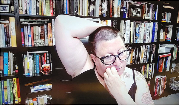 A photograph of a woman wearing glasses, her right arm behind her head while left hand is scratching her face, looking away from the camera and books on shelves in background