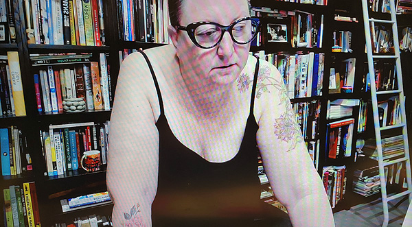 A photograph of a woman wearing glasses, looking away from the camera and books on shelves in background