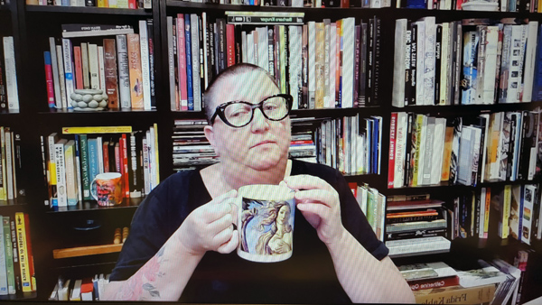 A photograph of a woman wearing glasses staring down, holding a coffee cup in both hands and books on shelves in background