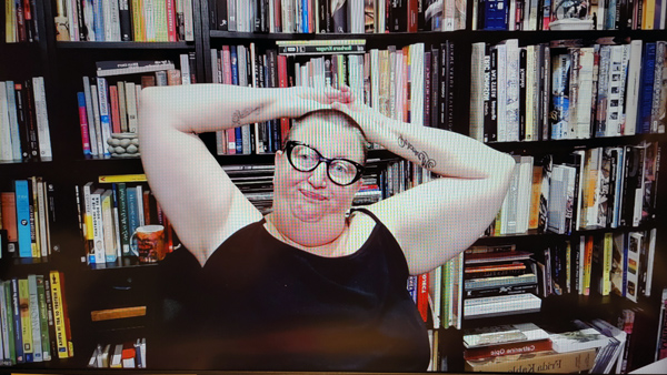 A photograph of a woman wearing glasses staring t the camera with with both hands folded resting on top of her head and books on shelves in background