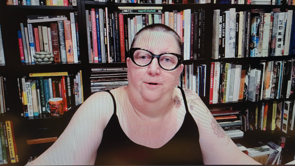 A photograph of a woman wearing glasses staring at the camera and books on shelves in background