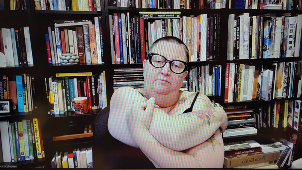 A photograph of a woman wearing glasses staring down with her arms folded in front of her and books on shelves in background