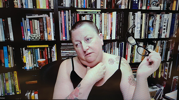 A photograph of a woman holding her glasses in her left hand with her right hand touching her neck staring off to her right and books on shelves in background