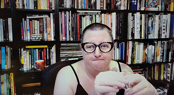 A photograph of a woman wearing glasses staring at the camera with her hands touching in the foreground and books on shelves in background