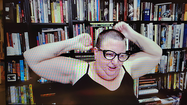 A photograph of a woman wearing glasses staring down with her arms folded behind her head and books on shelves in background