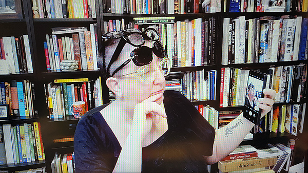 A photograph of a woman wearing four pairs of glasses on her face and head holding her cell phone in her left hand with her right hand touching her chin and books on shelves in background