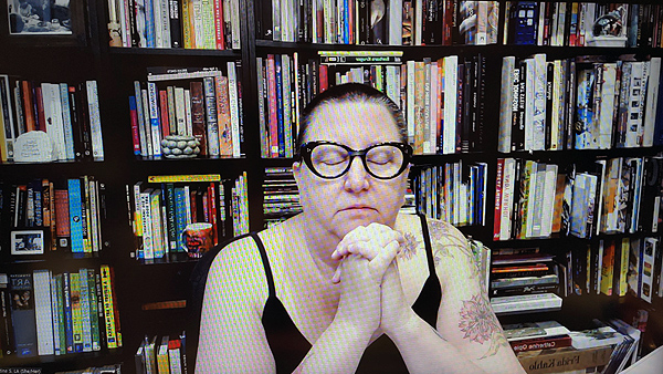 A photograph of a woman wearing glasses with her eyes closed with her hands folded resting on her chin and books on shelves in background