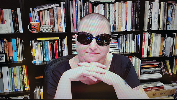 A photograph of a woman wearing sunglasses staring at the camera with her hands folded under her chin and books on shelves in background