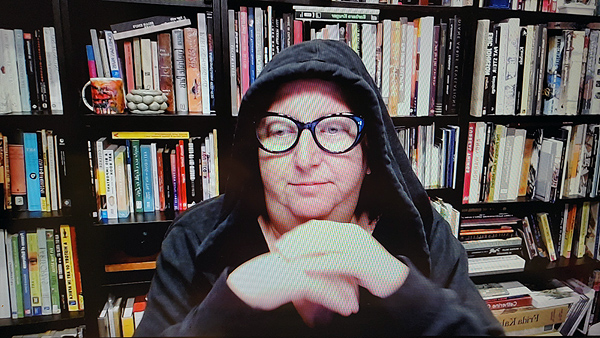 A photograph of a woman wearing a hoodies & glasses staring down to her right with her left hand on top of her right hand and books on shelves in background