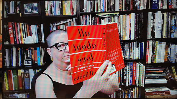 A photograph of a woman wearing glasses glancing to her right with her right hand holding a orange colored paperback book and books on shelves in background