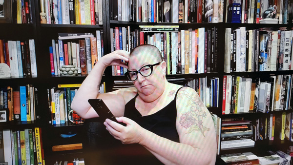 A photograph of a woman wearing glasses, her right hand on her head, looking at her phone in her left hand and books on shelves in background