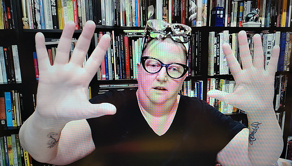 A photograph of a woman wearing glasses with two more pairs on her forehead, her her arms are raised with open hands facing the camera and books on shelves in background