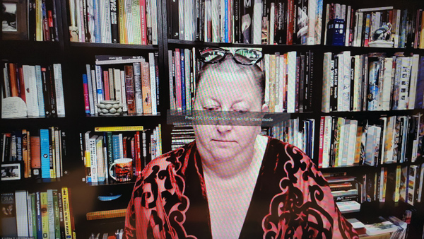 A photograph of a woman with a black strip across her eyes, glasses on her forehead, wearing a red & black robe and books on shelves in background