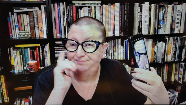 A photograph of a woman wearing glasses staring down with her right hand raised, her left hand holding a cell phone with her image on itand books on shelves in background