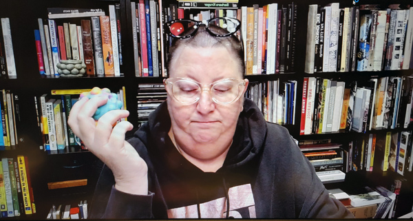 A photograph of a woman with glasses on her forehead staring down, holding a blue stress ball in her right hand and books on shelves in background