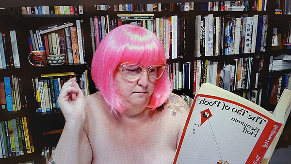 A photograph of a woman wearing glasses and a pink wig while reading a book and books on shelves in background