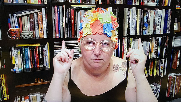 A photograph of a woman wearing glasses and a colorful swimming cap while both hands are pointing her forefingers upward and books on shelves in background