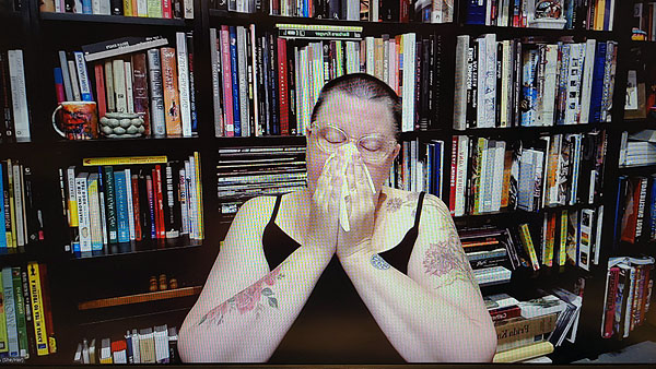 A photograph of a woman wearing glasses blowing her nose into a napkin and books on shelves in background