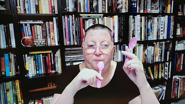 A photograph of a woman wearing glasses looking down while holding pink plastic spoons in each hand as well as behind both ears and books on shelves in background