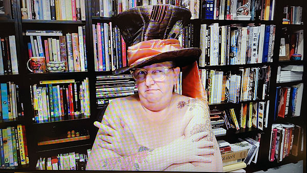 A photograph of a woman wearing glasses looking to the right wearing no shirt and a hat on her head and books on shelves in background