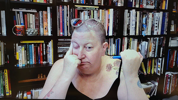 A photograph of a woman wearing glasses on top of her head while her right hand touches her face and a pen in her left hand and books on shelves in background
