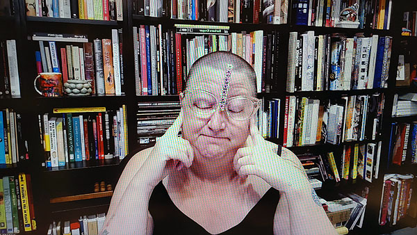A photograph of a woman wearing glasses, her eyes are closed with both her left and right forefingers touching the sides of her face with a vertical strip of small flower stickers on her forehead and books on shelves in background