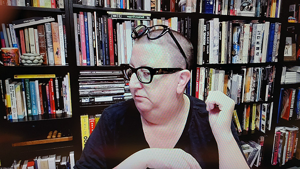 A photograph of a woman wearing glasses with another pair on top of her head staring to her right and books on shelves in background