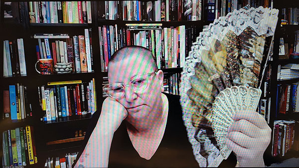 A photograph of a woman wearing glasses looking to the right holding a fan in her left hand while her right fist is resting on her face and books on shelves in background