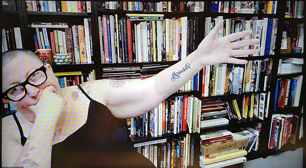 A photograph of a woman wearing glasses looking aat the camera leaning to her right while holding her left arm outstretched and books on shelves in background