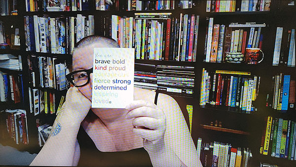 A photograph of a woman wearing glasses holding a card that is obstructing most of her face with words printed on it and books on shelves in background