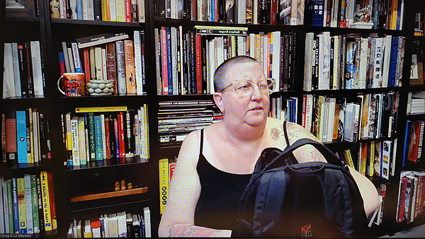 A photograph of a woman wearing glasses off to her right while digging in a blac purse with her left hand and books on shelves in background