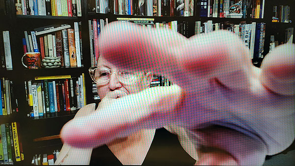 A photograph of a woman wearing glasses looking down, her right hand on her mouth while her left hand is reaching very closely to the camera and books on shelves in background