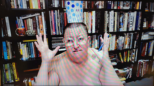 A photograph of a woman wearing glasses with no clothes on, a polka dot cup on her head with both hands open on either side of her head and books on shelves in background